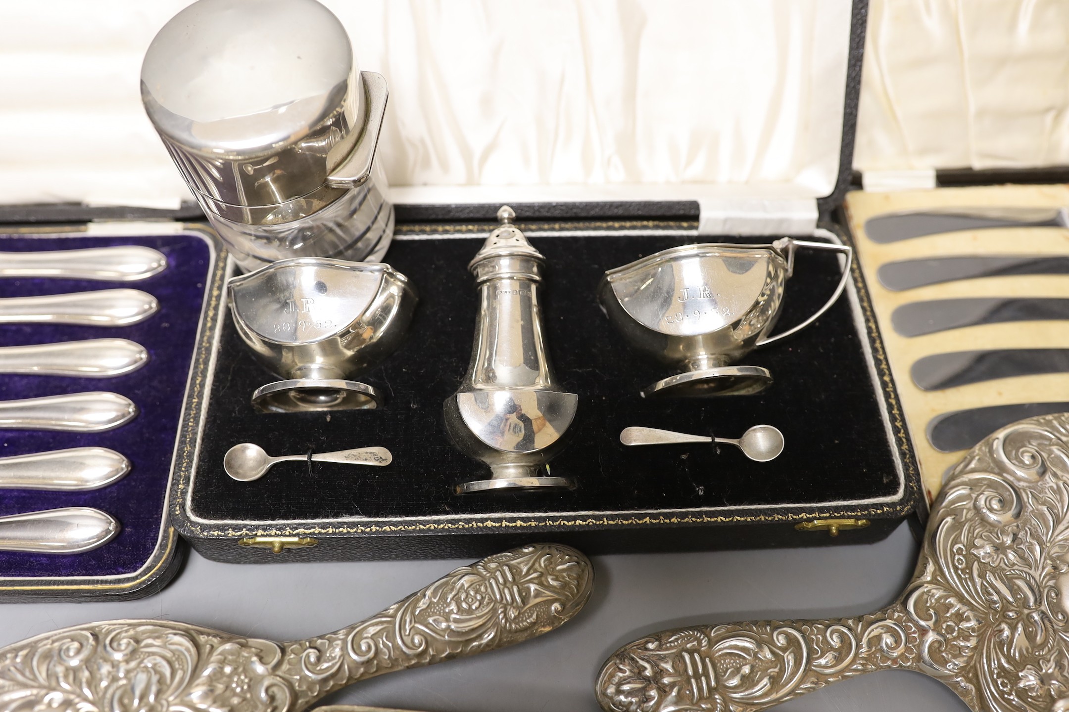 A cased 1950's silver three piece condiment set, two cased sets of six silver handled tea knives, a silver mounted glass salts bottle, silver mounted hand mirror and two brushes.